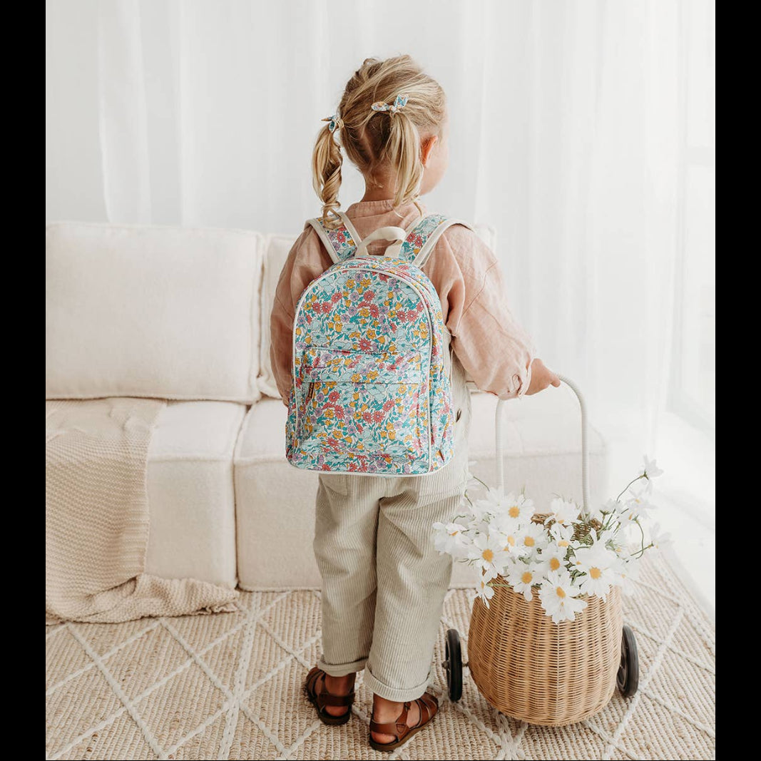 A child standing by a couch, wearing a multicolored floral backpack with adjustable straps.