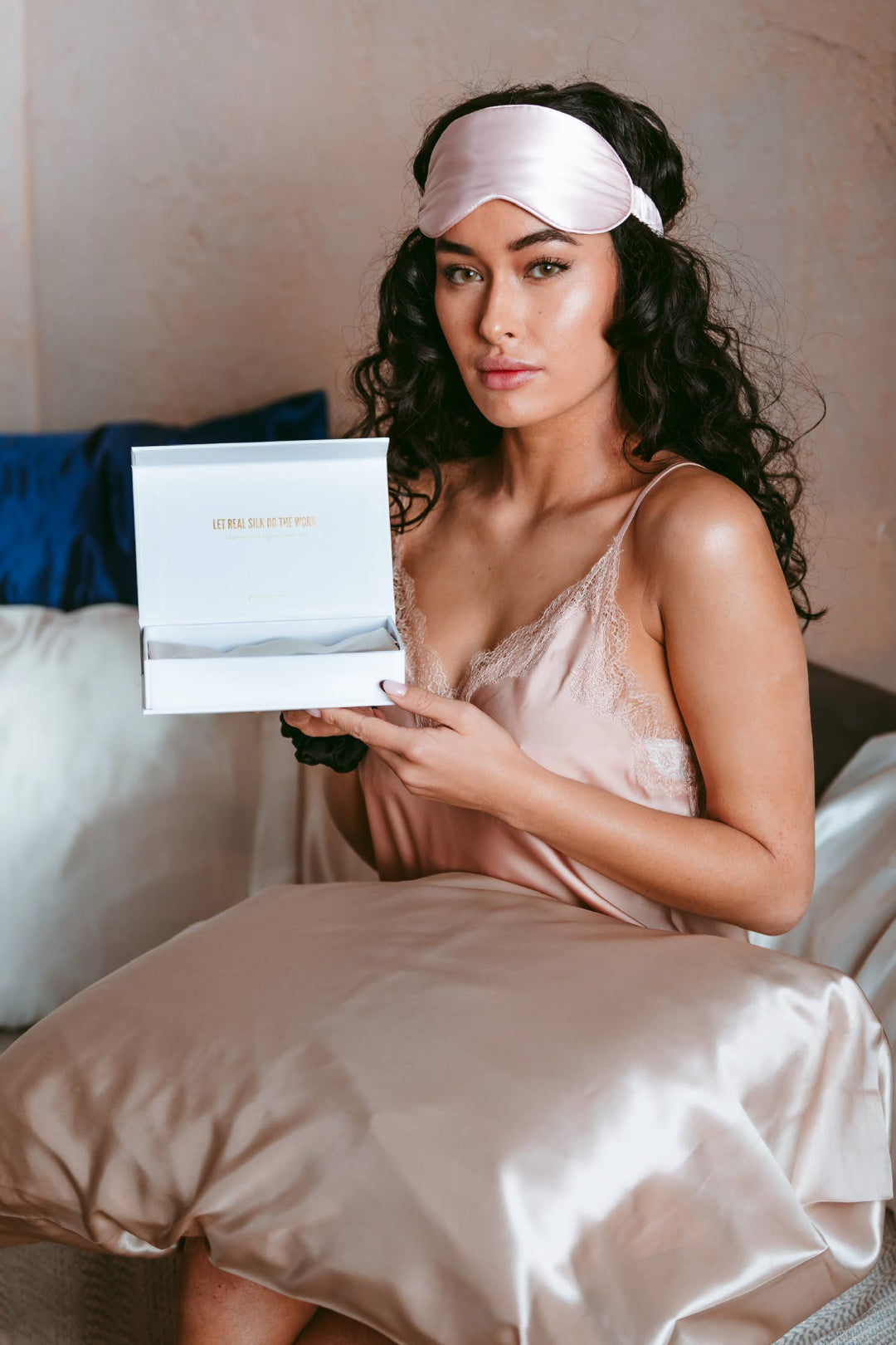 Woman wearing a silk eye mask and holding a product box in bed.
