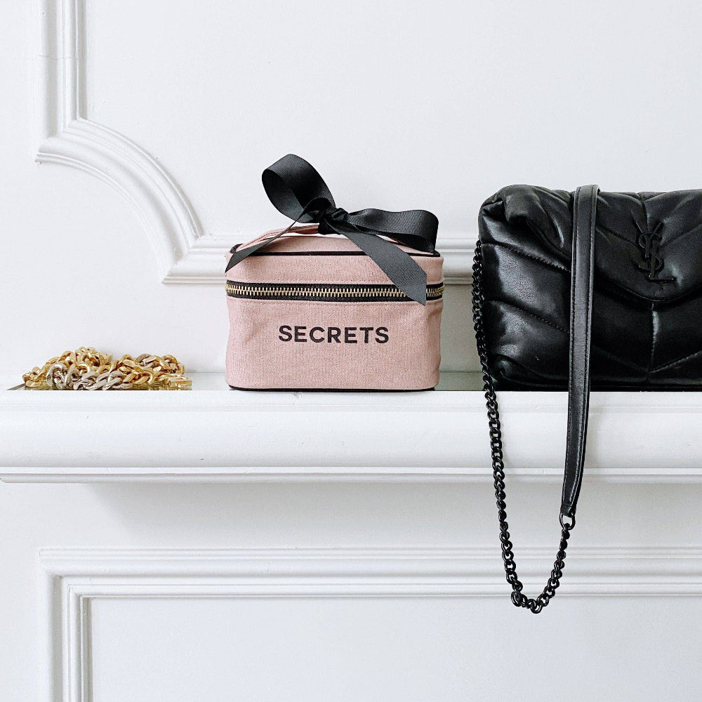 Pink cosmetic bag with 'SECRETS' text and black bow, next to a black handbag on a white surface.