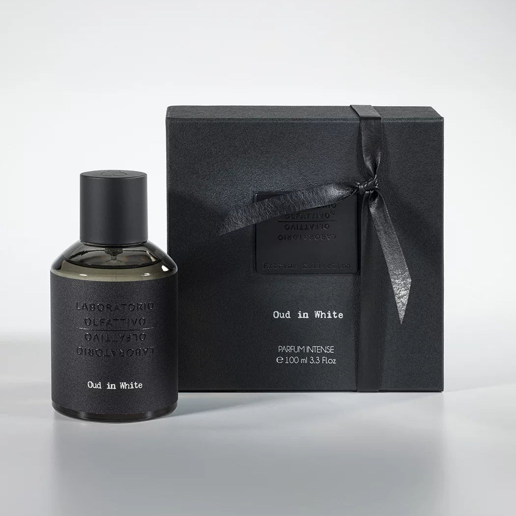 A bottle of Oud in White 100ml Eau de Parfum next to its black box packaging adorned with a ribbon.