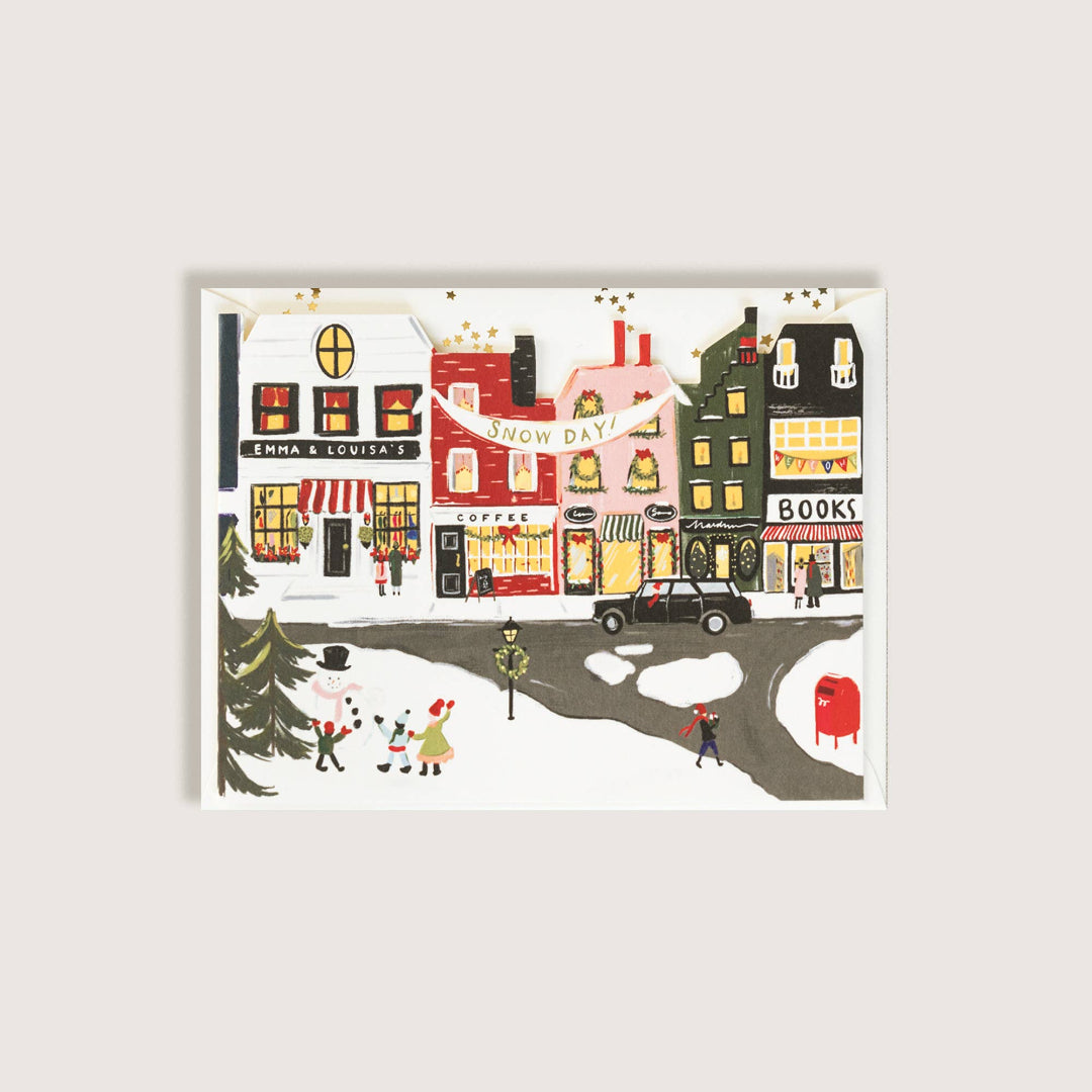 A greeting card depicting a whimsical winter street scene with colorful storefronts, snow, tiny people, and a red mailbox, shown against a light gray background.