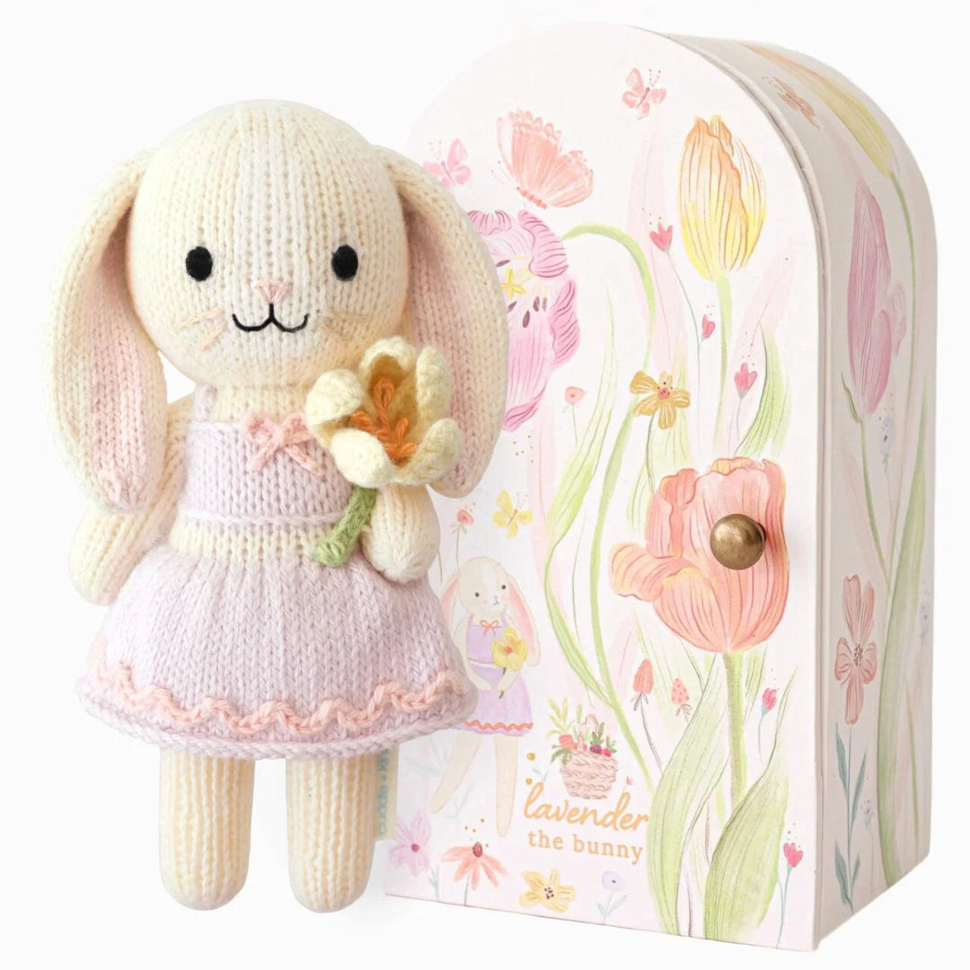 A knitted bunny doll wearing a pale pink dress stands beside a decorative illustrated box labeled 'lavender the bunny'