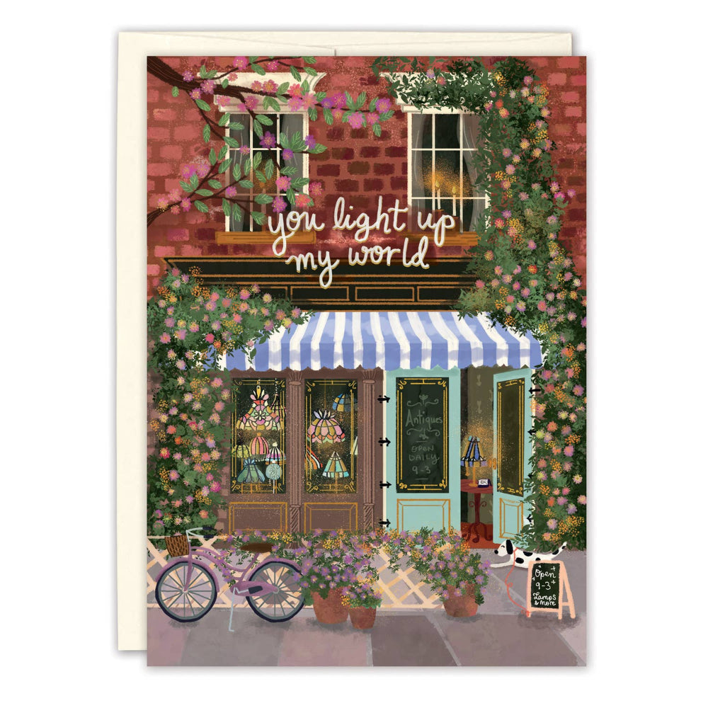 Illustrated greeting card featuring a storefront with the phrase 'You light up my world', gold foil details, and a bicycle in the foreground.