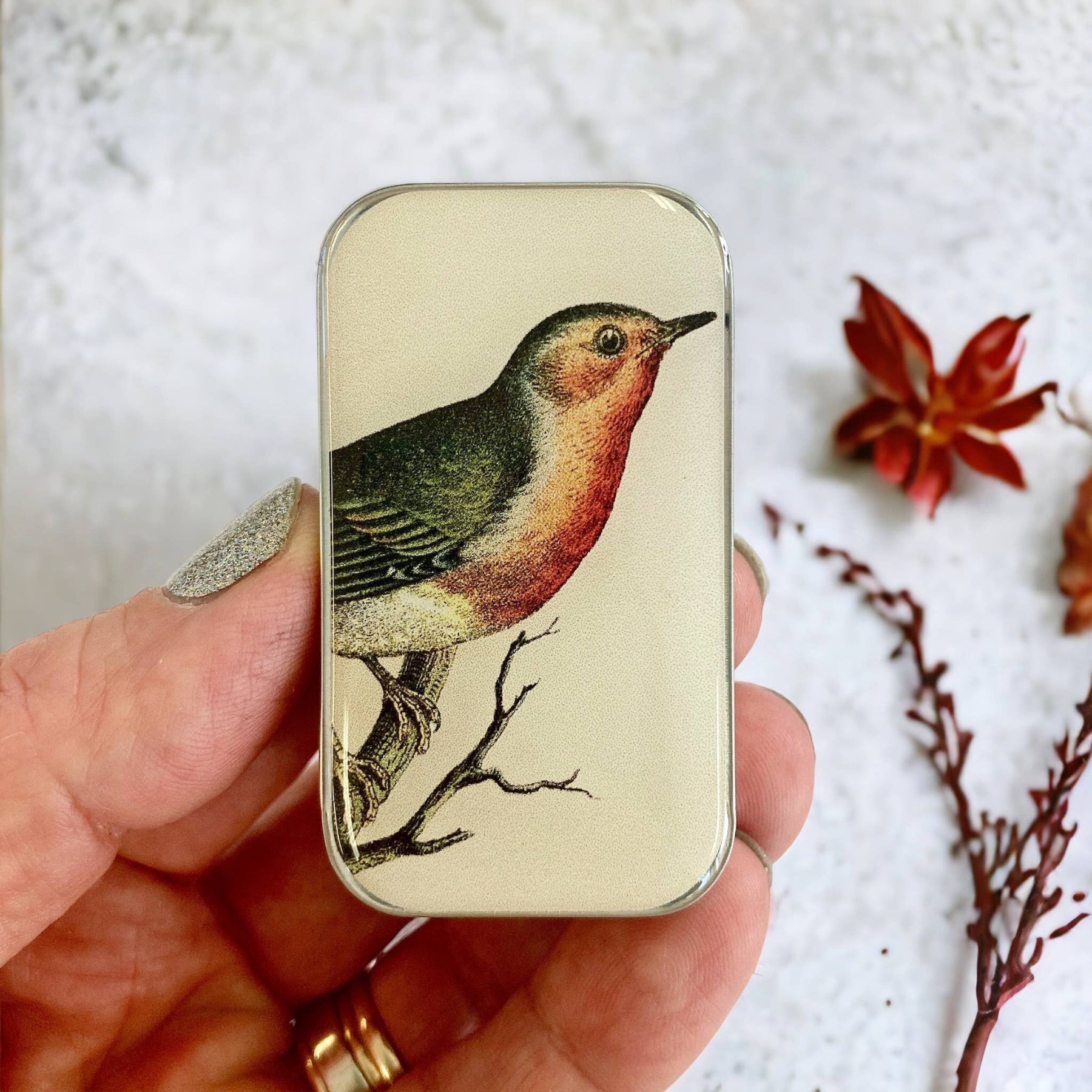 A hand holding a small tin with a vintage robin illustration on the lid, with autumnal decorations in the background.