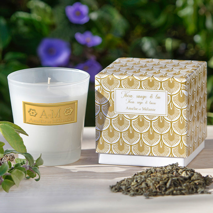 A clear scented candle with a label that reads 'Theia, Sage & Linen' placed next to its decorative box with a floral pattern. The candle and box are set on a wooden surface with some dried plant leaves nearby.
