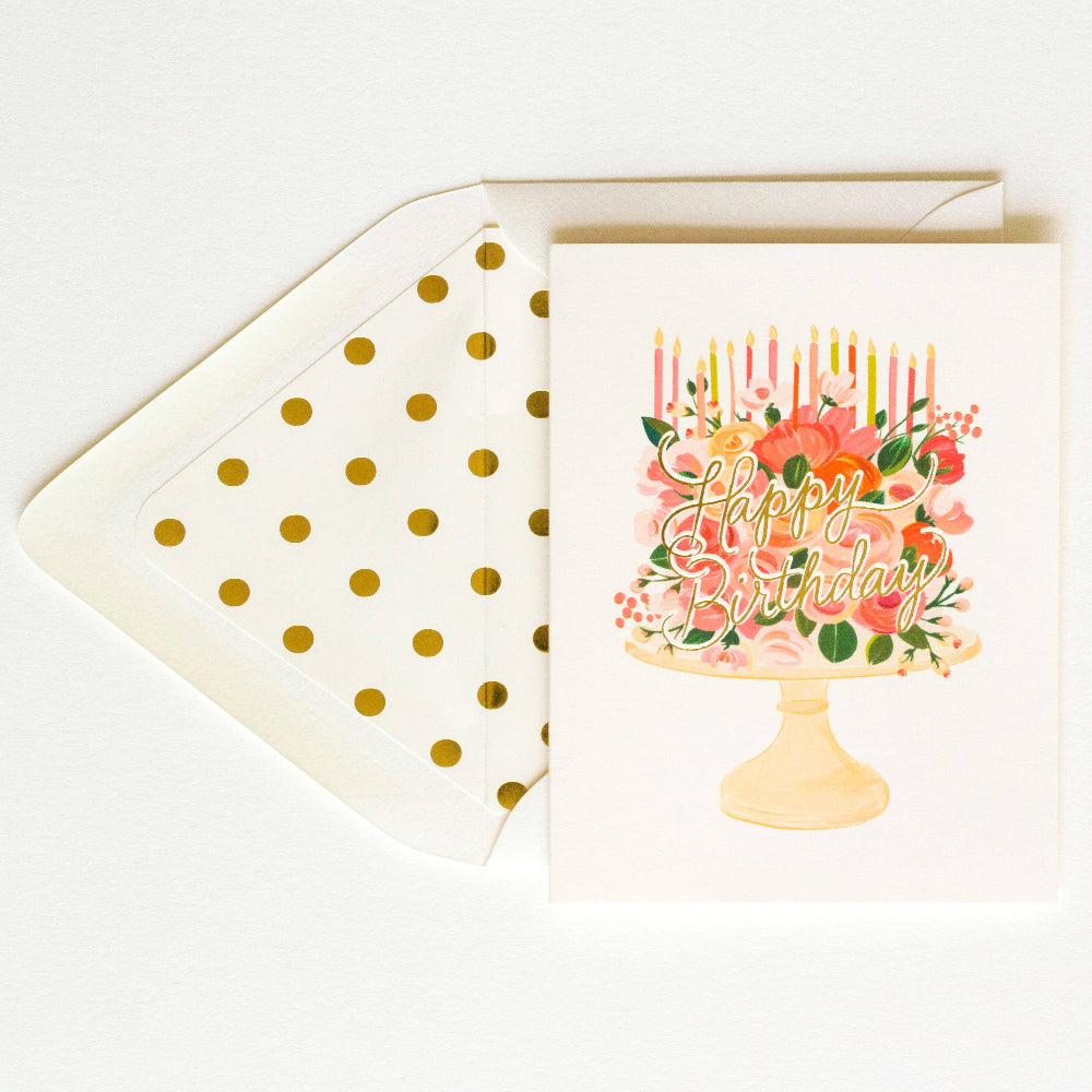 Greeting card with a birthday cake design, featuring flowers and candles, on a white background with gold dotted lines and a cream envelope.