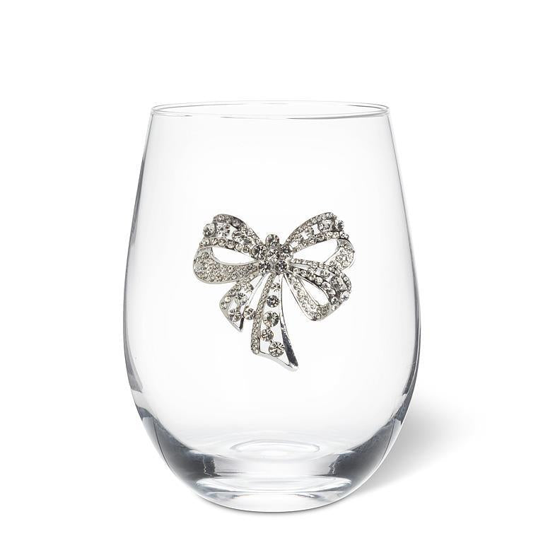 A clear stemless wine glass featuring a decorative silver bow embellished with sparkling clear rhinestones, set against a plain white background.