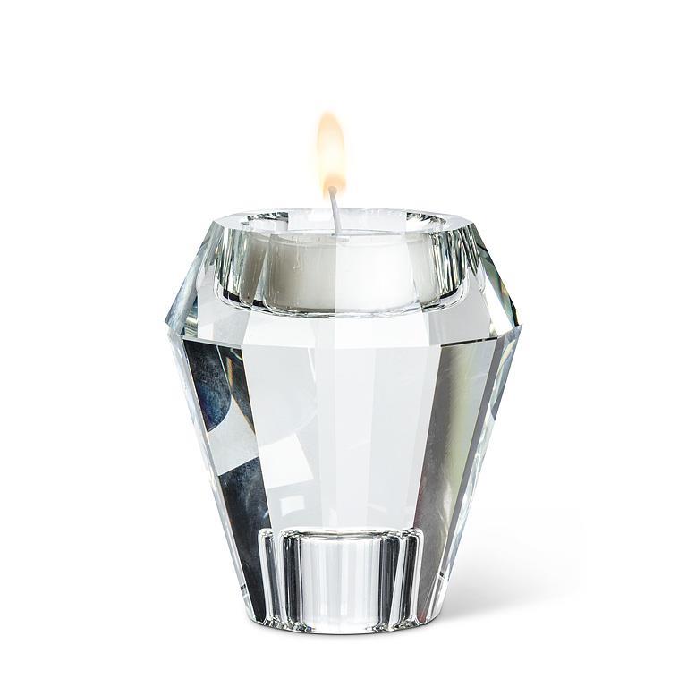 A clear faceted glass candle holder shaped like a diamond, holding a white tea light candle with a small flame, photographed on a white background.