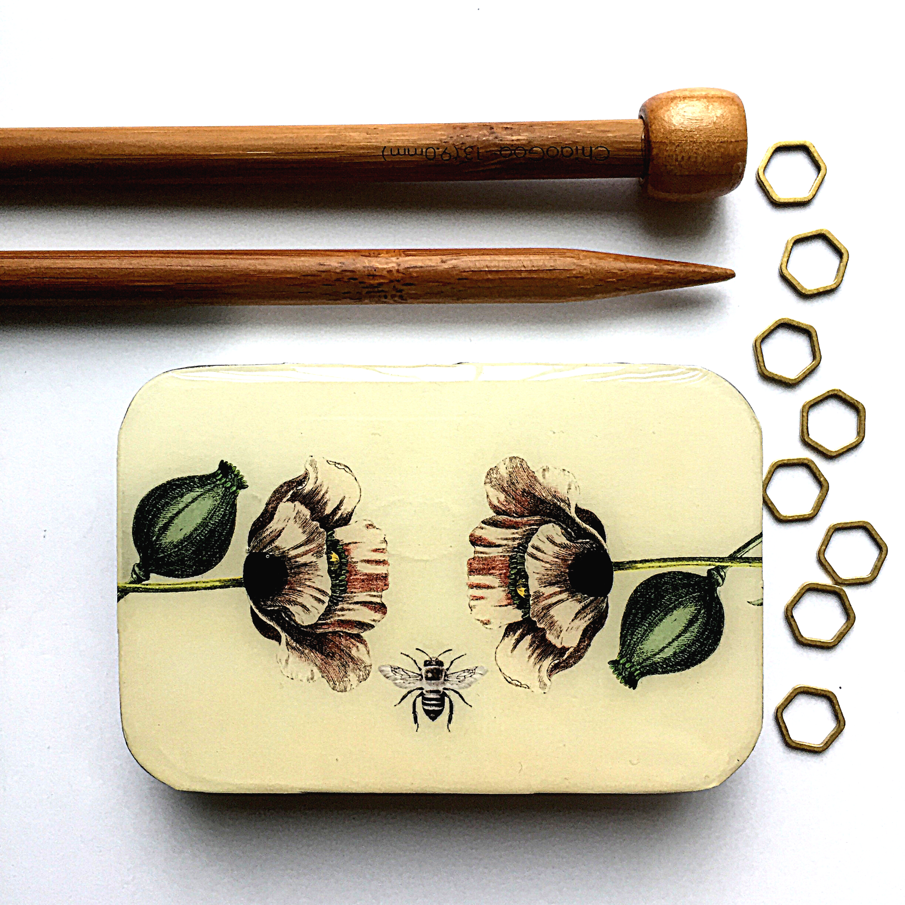 Decorative tin with floral design, wooden knitting needles, and metal hexagonal shapes on a white background