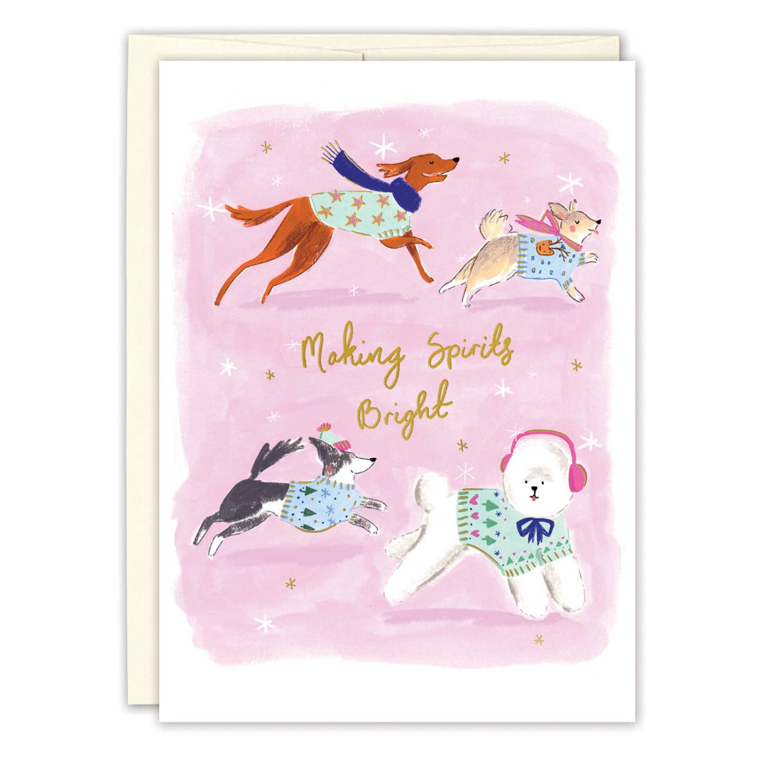 A greeting card with a watercolor illustration of four dogs wearing knit sweaters on a pink background, with the text 'Making Spirits Bright' in gold script. A cream envelope is visible behind the card.
