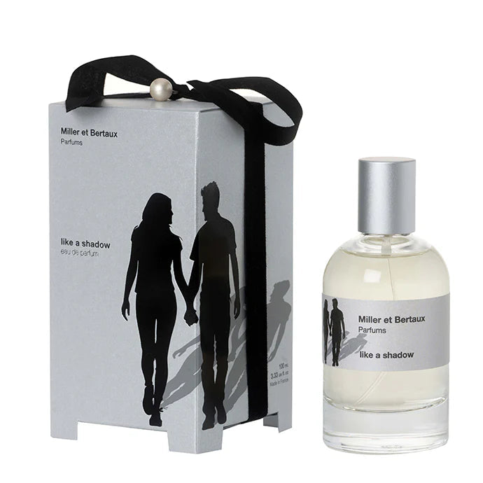 Miller et Bertaux Perfums 'like a shadow' eau de parfum, featuring a silver-gray gift box with a black ribbon and silhouette graphic next to a clear glass bottle with a silver cap.